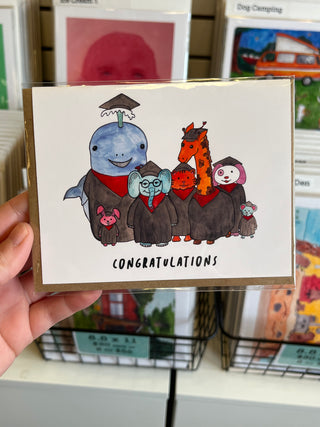 Hand holding a card with cartoon characters and 'Congratulations' text, with a bookshelf in the background.